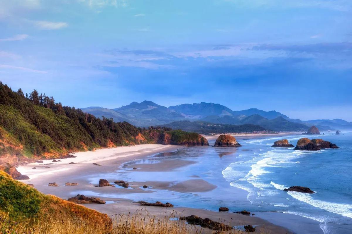 This image captures a tranquil stretch of sandy beach along a rocky coastline, with gentle waves lapping the shore and distant mountains rising in the background. The scene suggests opportunities for hiking, beachcombing, and peaceful seaside walks at dawn or dusk.