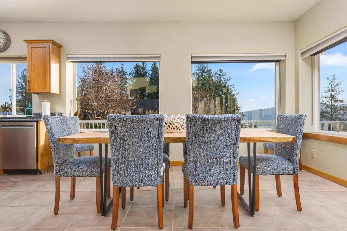 The dining area is anchored by a central table, with large windows in the background offering glimpses of outdoor scenery and letting sunlight illuminate the space. The kitchen area is visible, creating a sense of openness and connection among the main living zones.