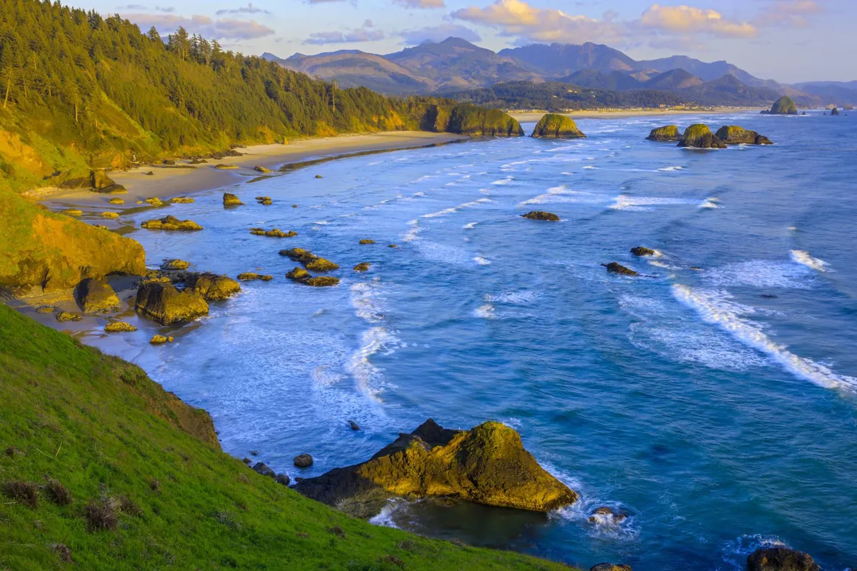 A sweeping view of the Oregon Coast, featuring gentle waves rolling onto a sandy beach framed by dramatic rock formations, coastal forest, and distant mountains. The scene captures the natural beauty and adventure opportunities that families can enjoy, such as beachcombing, tidepooling, and exploring hidden coves.