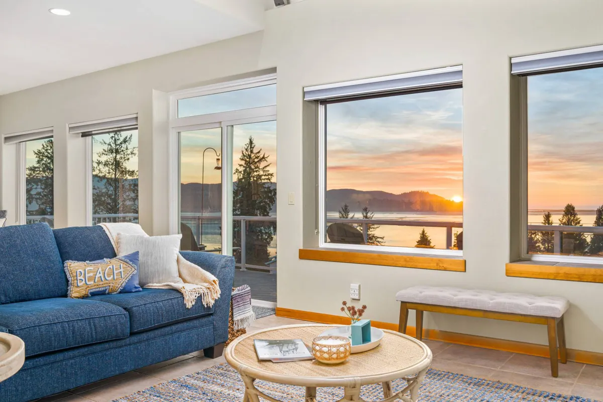 An inviting living area featuring a blue sofa, a coffee table, and large windows that frame a sunset view. The setting is cozy yet stylish, with a decorative pillow reading 'BEACH' enhancing the coastal ambiance. The composition blends comfort with scenic vistas.