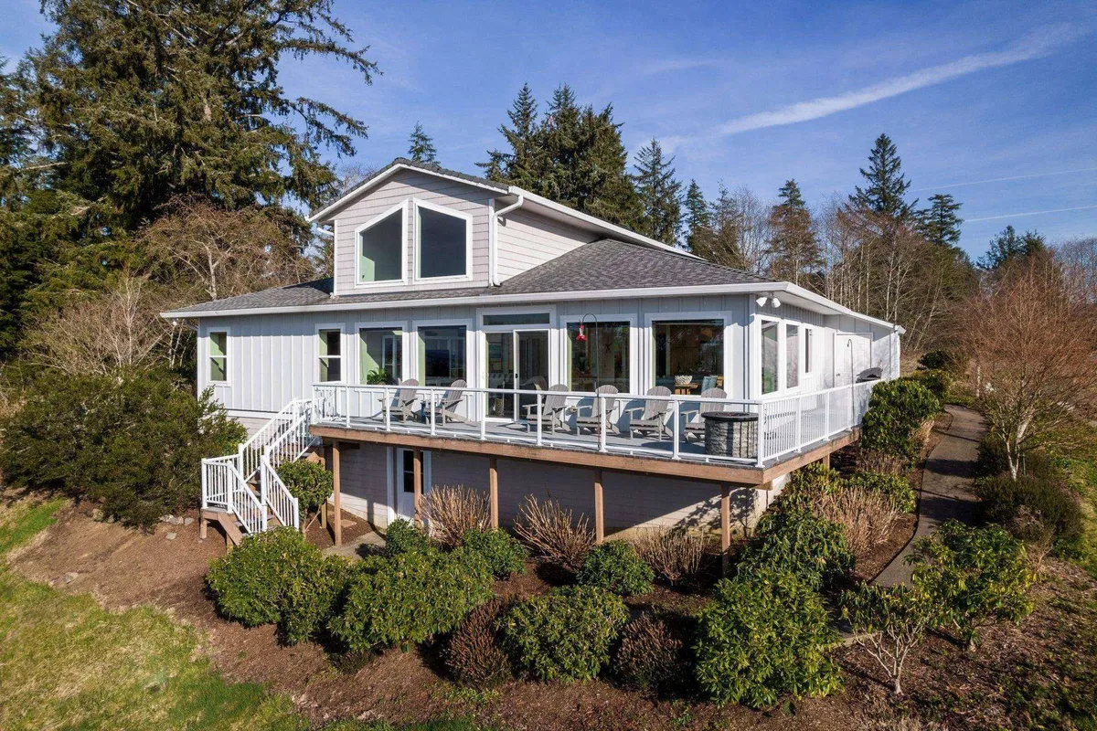 A large, modern deck attached to the house, framed by lush greenery and large windows, inviting outdoor dining, relaxation, and enjoyment of the sunset or morning light. The seamless transition between indoor and outdoor spaces is clearly visible.
