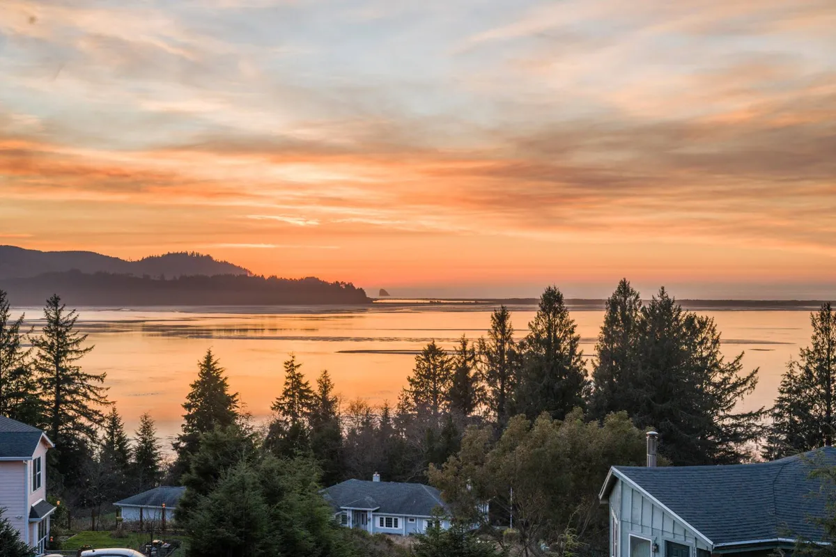 This view captures a breathtaking sunset over the water, silhouetted trees, and distant hills in the background. The mood is calm and serene, ideal for relaxing after a workday or appreciating the coastal landscape.