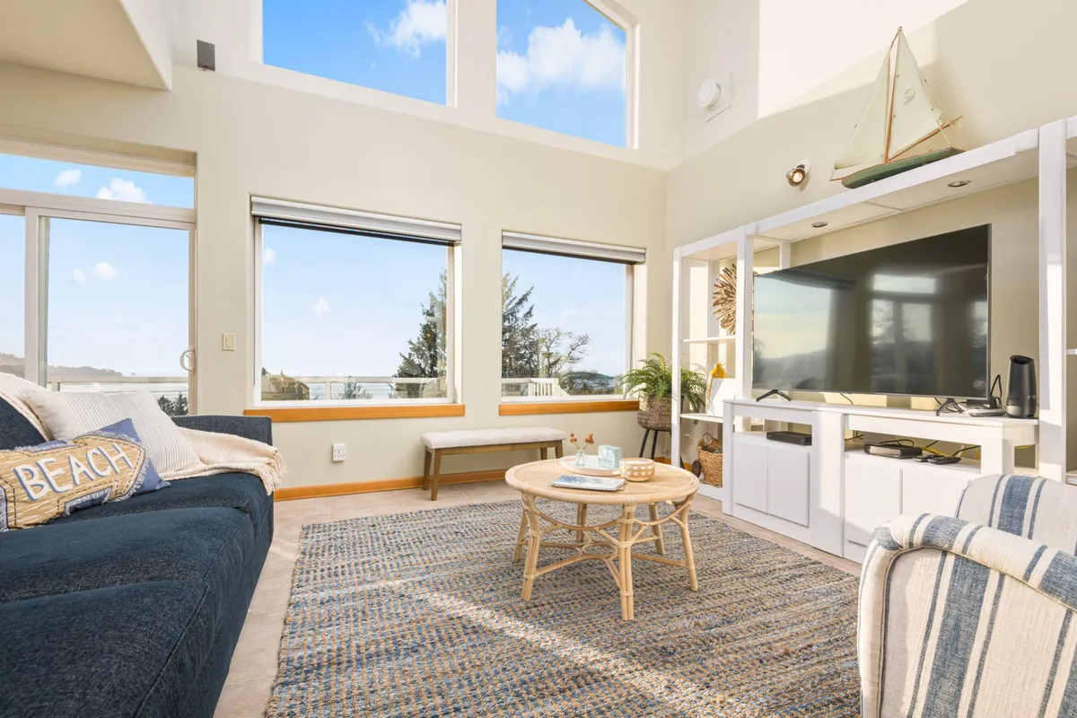 This image showcases a spacious, open living area featuring a comfortable blue sofa positioned to take in the abundant natural light pouring through floor-to-ceiling windows. A coffee table and a television unit complete the scene, with the arrangement oriented toward panoramic views outside, evoking a sense of both relaxation and connection to the surrounding seascape.