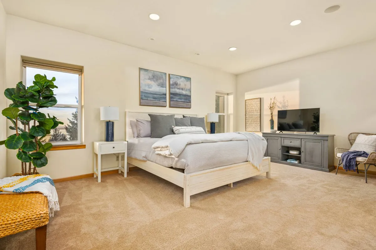 A serene bedroom with gray bedding on a large bed, two bedside lamps, a large window providing plenty of natural light, a wall-mounted television, and tasteful artwork. The setting exudes comfort and privacy, making it suitable for families or guests.