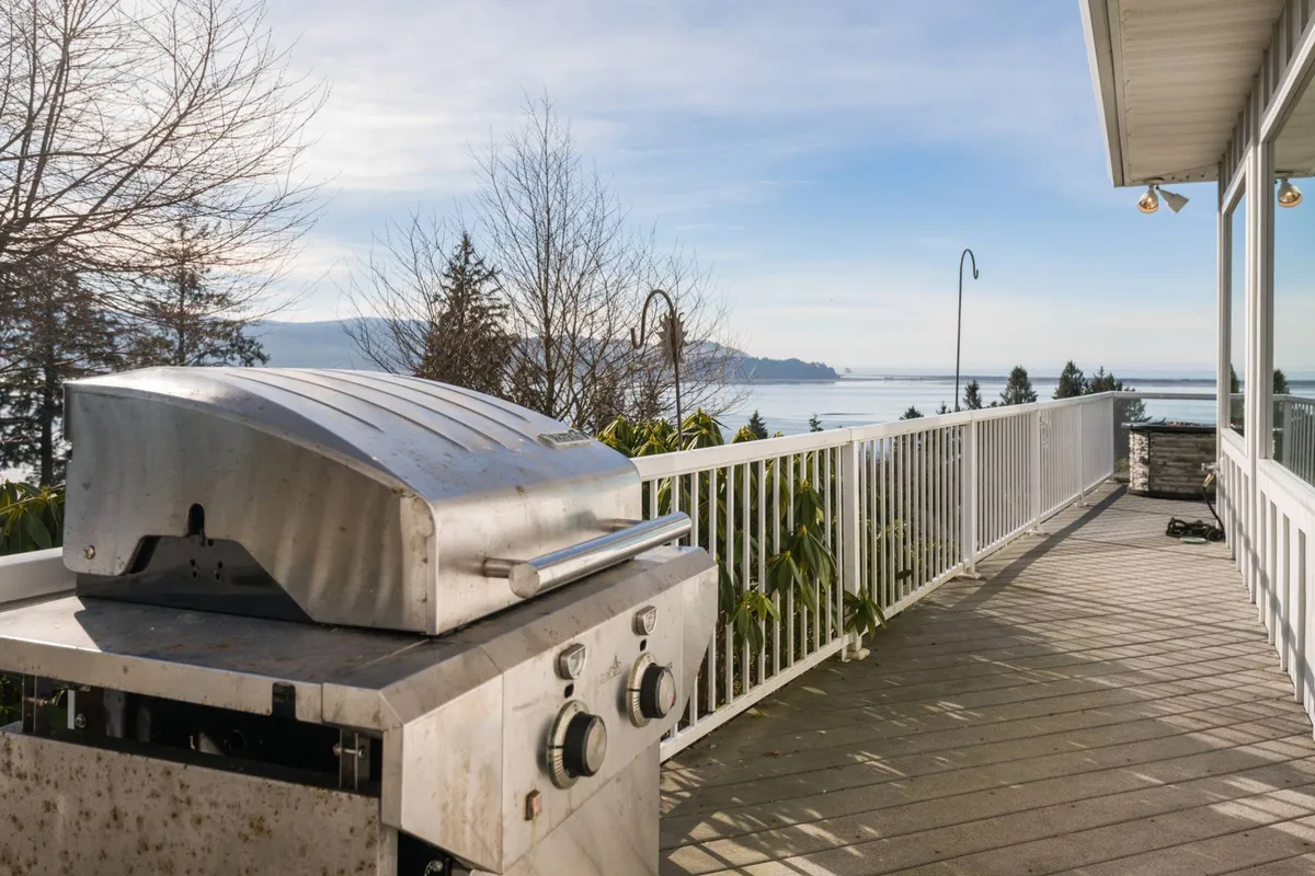 An inviting exterior view from a deck or patio overlooking the water, with a grill, railing, and trees in the foreground. The peaceful waterfront setting invites relaxation and outdoor gatherings, with ample space for enjoying the fresh air.
