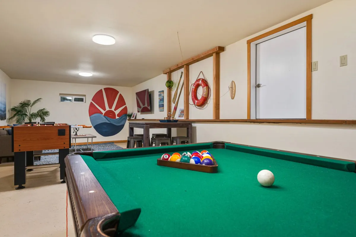 This image showcases a spacious game room featuring both a pool table and a foosball table, with a sunset mural and a dining table in the background. The layout suggests ample space for group activities and fun gatherings.