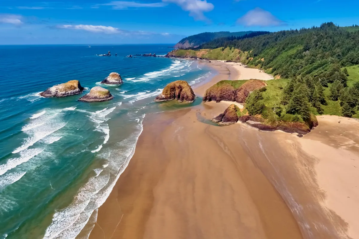 A picturesque view of a sandy beach framed by dramatic rock formations and a forested hillside, with gentle waves rolling in from the ocean. The setting is ideal for swimming, sunbathing, hiking, and beachcombing, all in a tranquil environment.