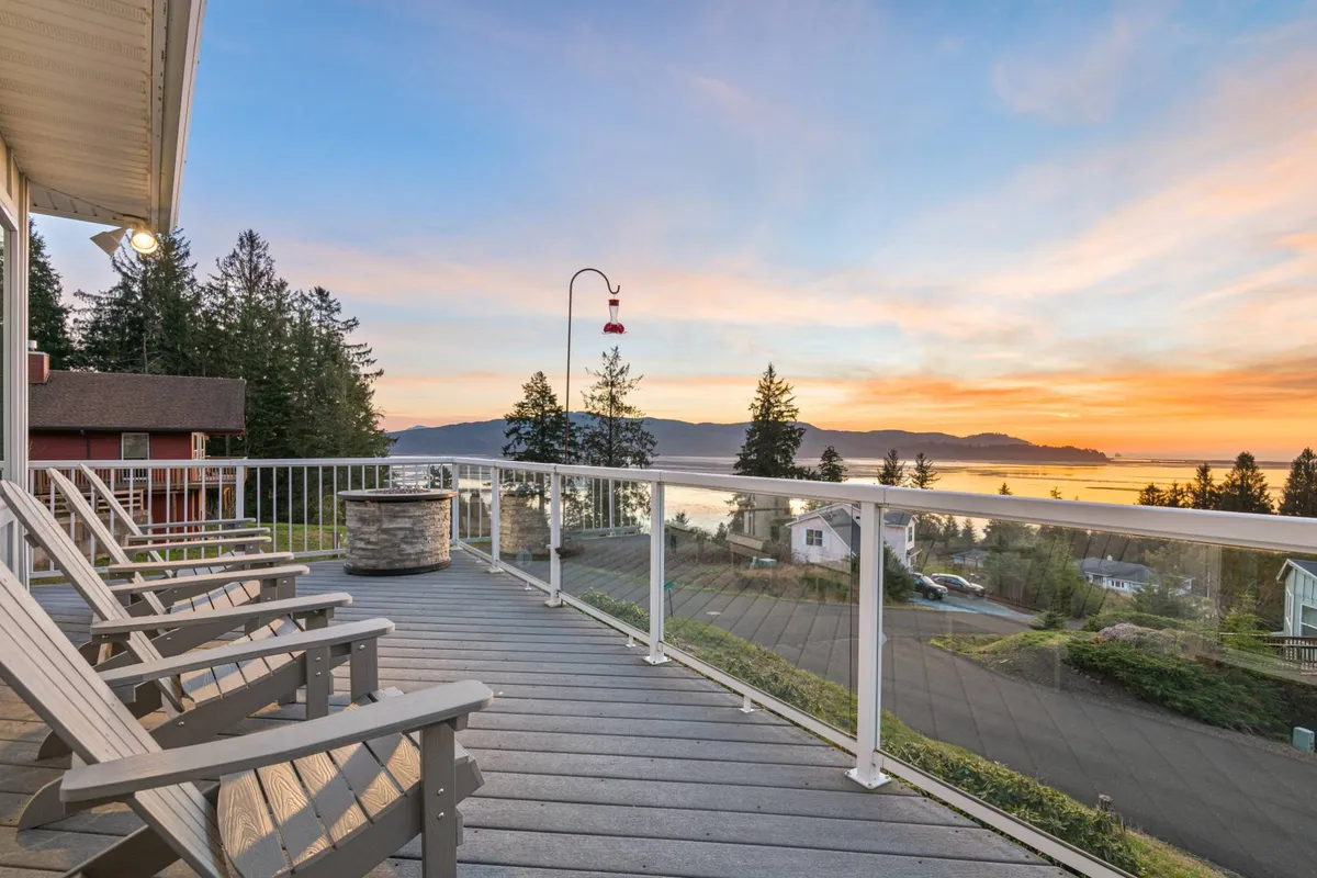 The image showcases a serene outdoor seating area set atop a hillside, offering a front-row view of a glowing sunset over Tillamook Bay and the vast Pacific Ocean. Surrounded by lush trees, the inviting deck is perfect for relaxing, socializing, or simply soaking up the tranquil coastal beauty.