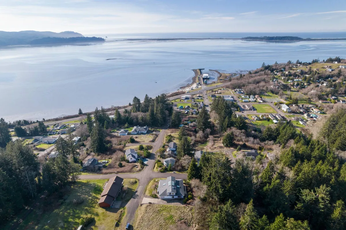 A tranquil waterfront scene showcasing homes nestled among trees, with a view of calm water, forested hills, and local roads. The setting hints at nearby community parks and easy access to both nature and local amenities.