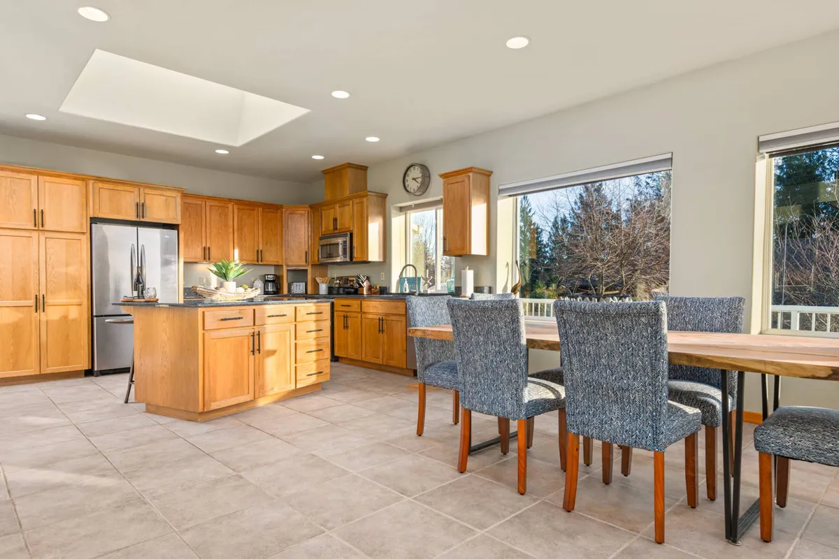 A warm, welcoming space with a well-equipped kitchen island, wooden cabinetry, a dining table, and large windows. The arrangement flows seamlessly, visually connecting the kitchen and dining areas, while the windows flood the space with natural light and frame the picturesque surroundings.